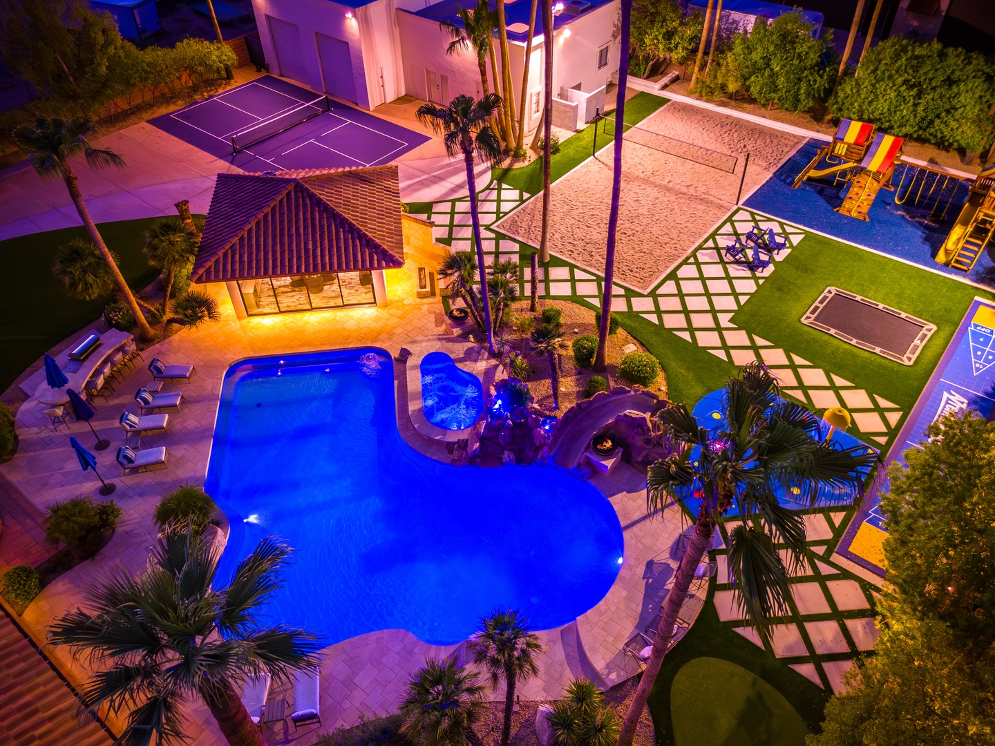 Aerial view of the property at night showcasing an illuminated pool, tennis court, children's playground, and tropical landscaping across expansive grounds.