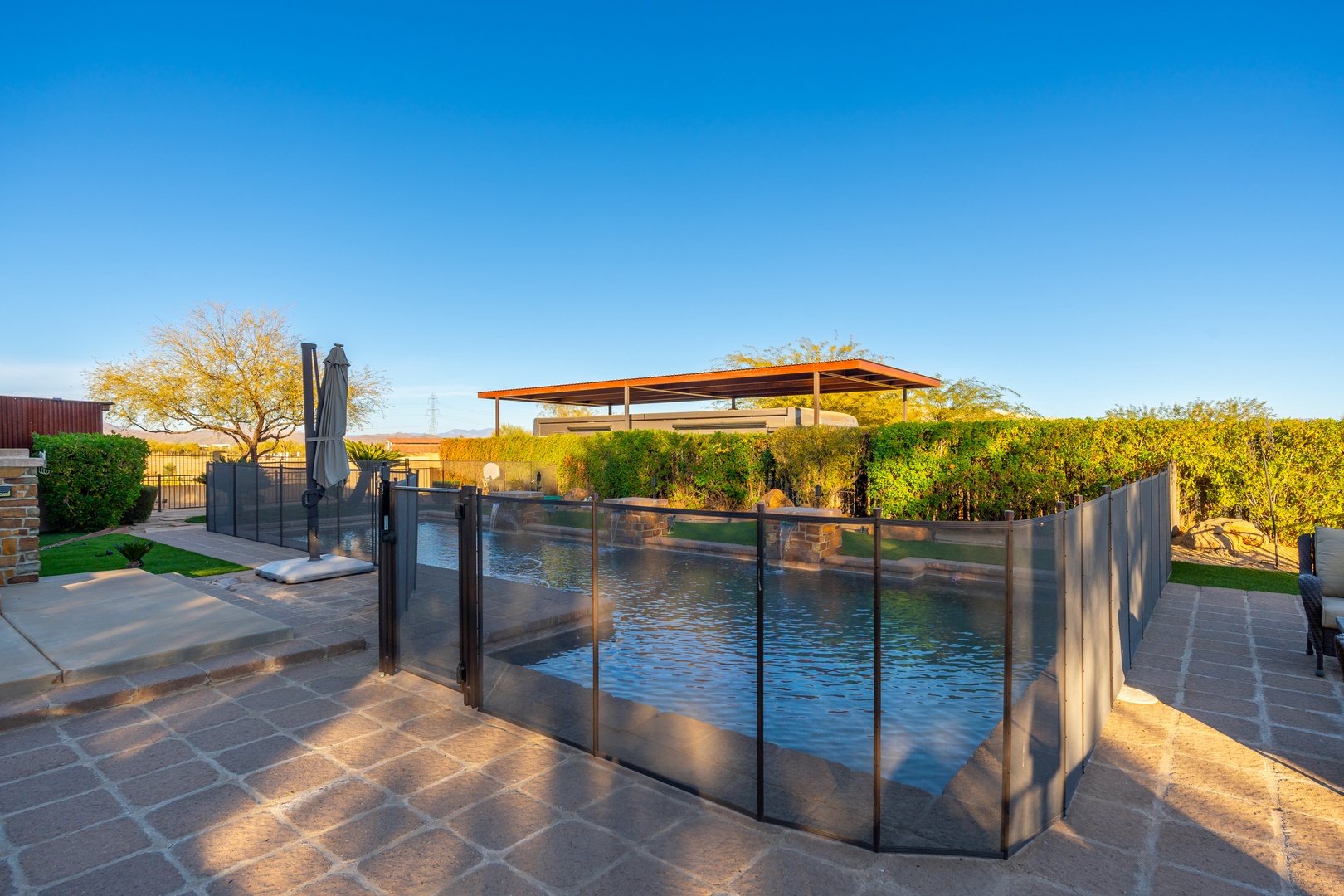 A sparkling pool with safety fencing, surrounded by lush landscaping and a shaded cabana, invites relaxation under clear blue skies.