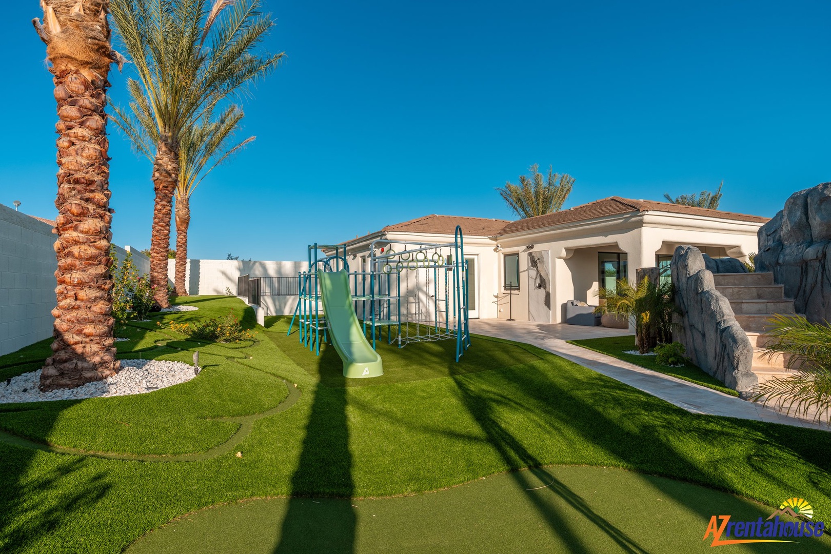 Beautiful property exterior featuring manicured gardens, towering palm trees, and playground area under clear blue skies.