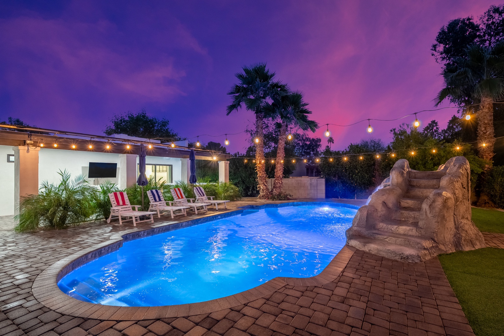 The property features a stunning twilight scene with a pool, waterslide, string lights, and surrounding palms creating an inviting outdoor entertainment space.
