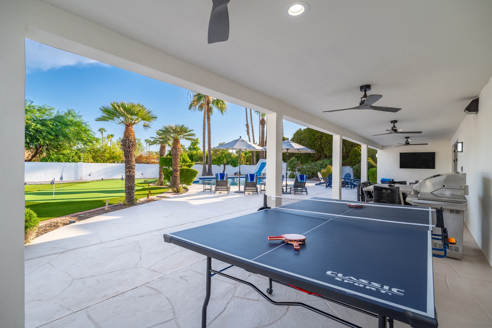 Step into your covered patio retreat where ping pong awaits, ceiling fans keep you cool, and lush palms frame your private backyard oasis with lounge chairs and dining under sunny skies.