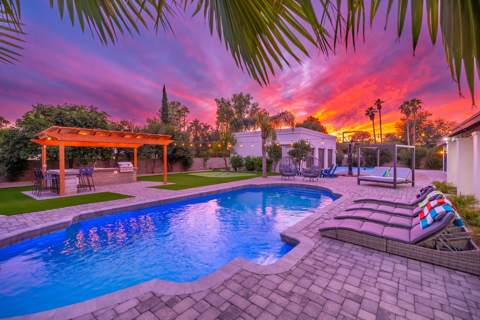A dramatic sunset paints the sky over this private resort-style backyard featuring a sparkling pool, lounge chairs with colorful pillows, covered patio dining, and lush tropical palms framing the scene.