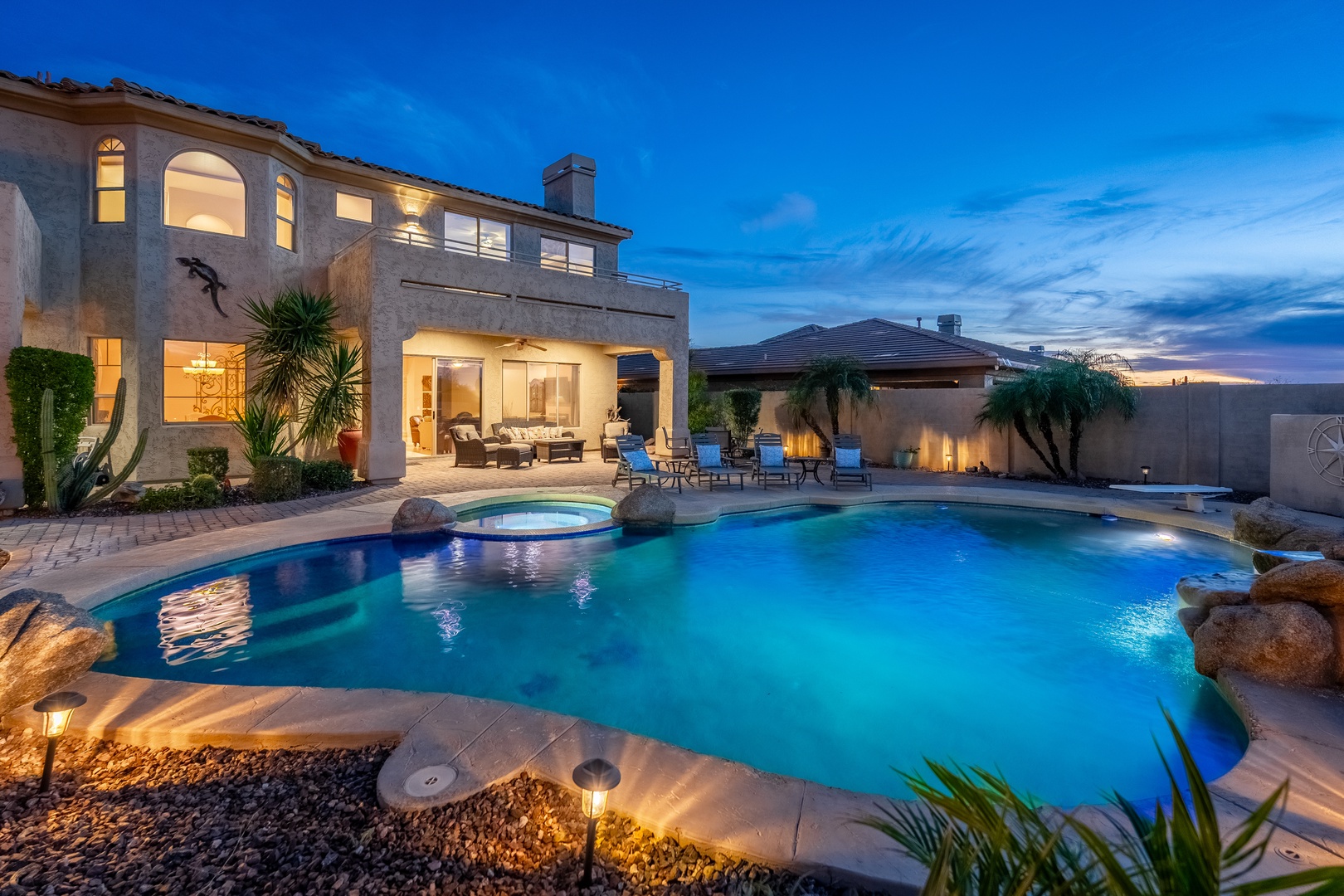 An elegant two-story property showcasing its inviting pool and hot tub at twilight, with warm interior lighting and a spacious patio perfect for evening gatherings.