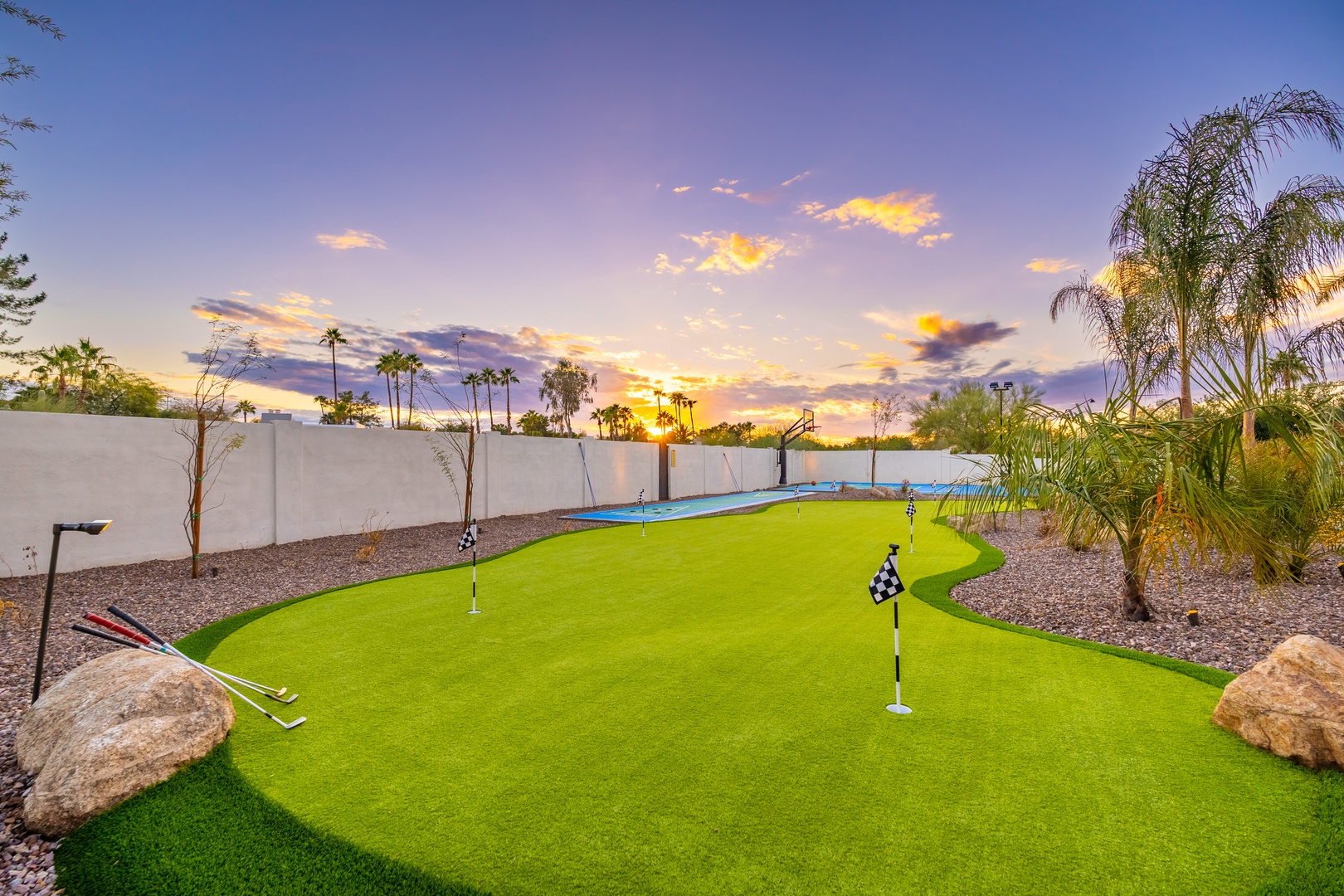 A private backyard features a custom putting green and swimming pool, set beneath palm trees with desert landscaping and a vibrant sunset backdrop.