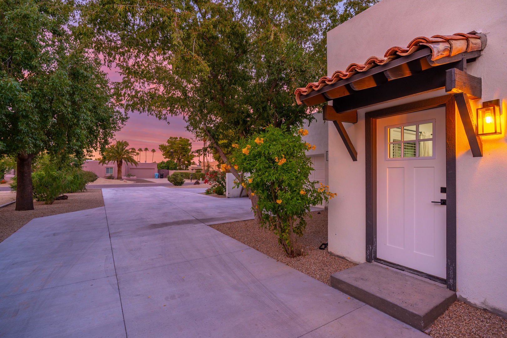 The charming entryway features Spanish-tile accents and warm lighting against a stunning twilight sky, setting the tone for a welcoming desert retreat with mature trees and landscaping nearby.