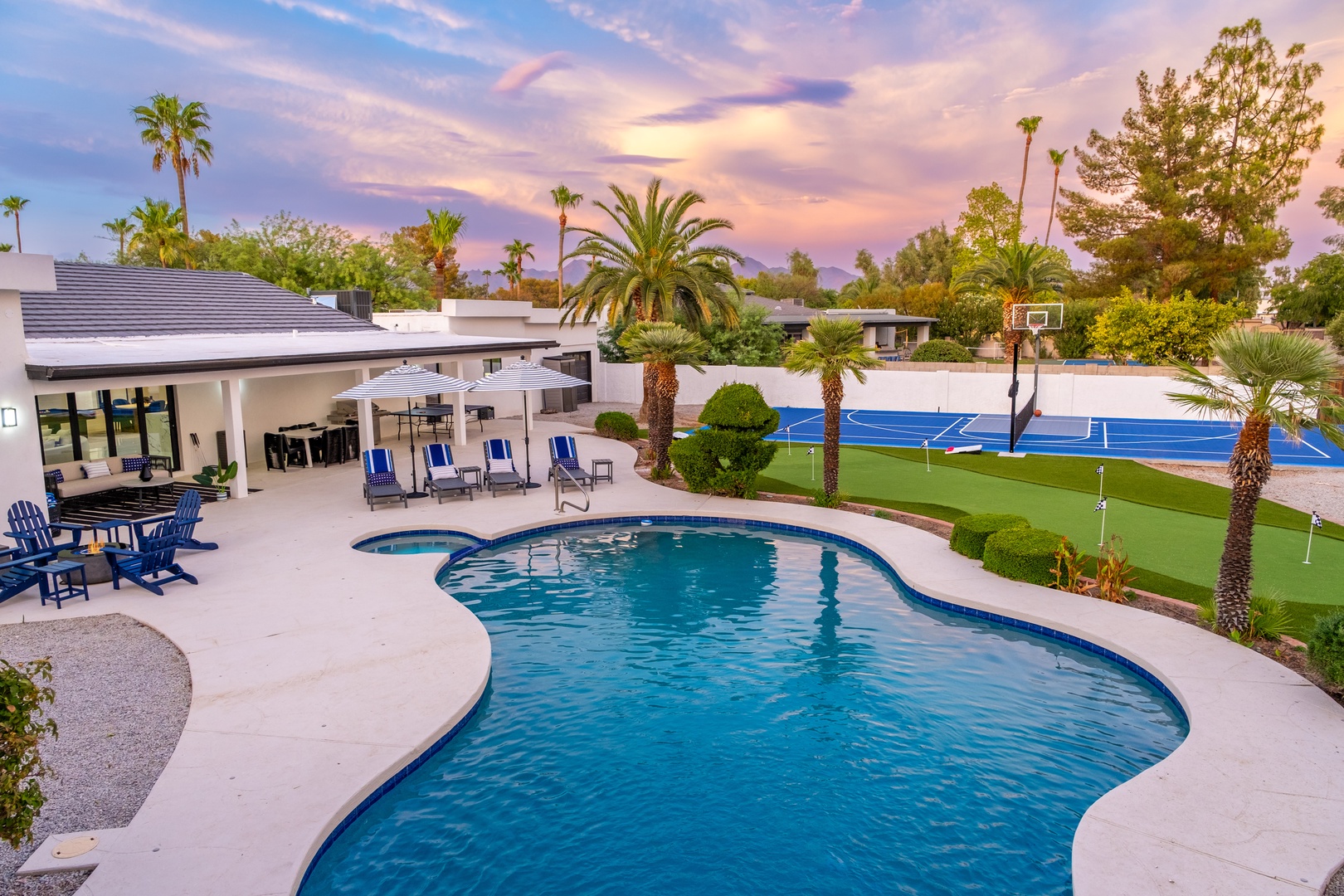 A stunning desert oasis featuring a resort-style pool, private basketball court, and multiple lounging areas beneath swaying palms, framed by a pastel twilight sky—an ideal setting for outdoor living and entertainment.