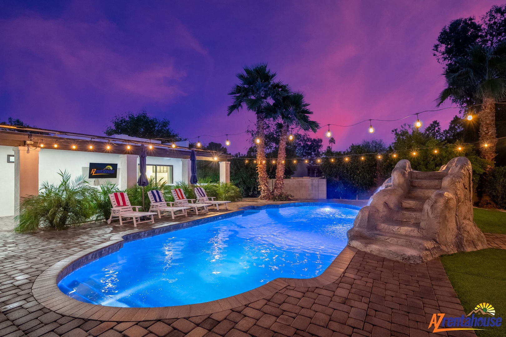 Stunning twilight scene featuring a glowing pool with rock slide beneath purple-pink skies and twinkling string lights.