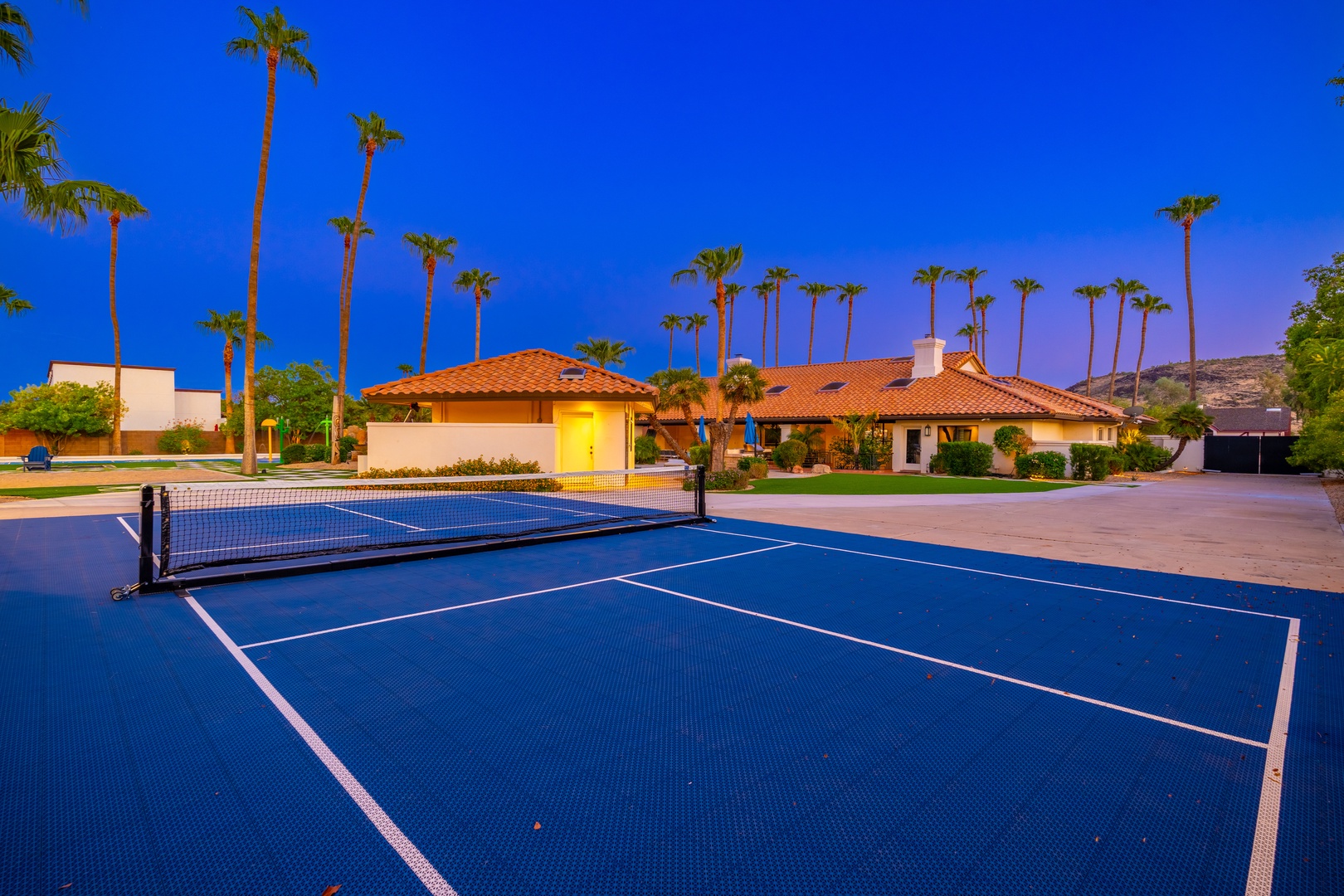 This property features a pristine private tennis court set against swaying palms and a desert hillside backdrop, captured at twilight.