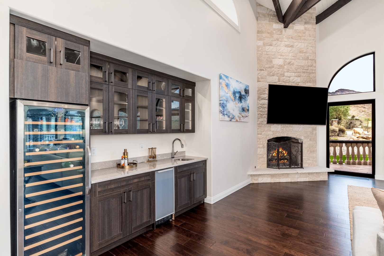 A modern wet bar featuring a sleek wine fridge and glass-front cabinetry anchors this stunning space, where exposed beams and a stone fireplace create an inviting atmosphere for your gatherings.
