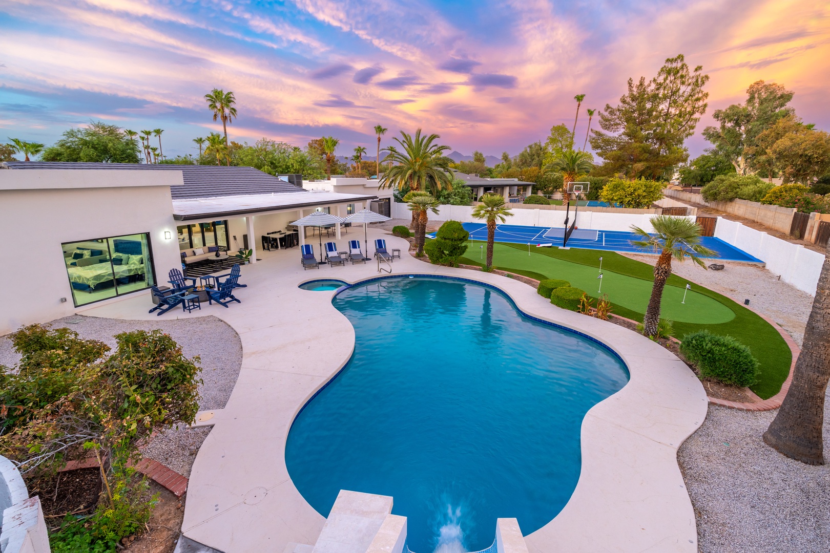 An elevated view captures a modern vacation rental property featuring an inviting pool, miniature golf course, and spacious patio beneath a stunning painted sky—a private oasis awaits.