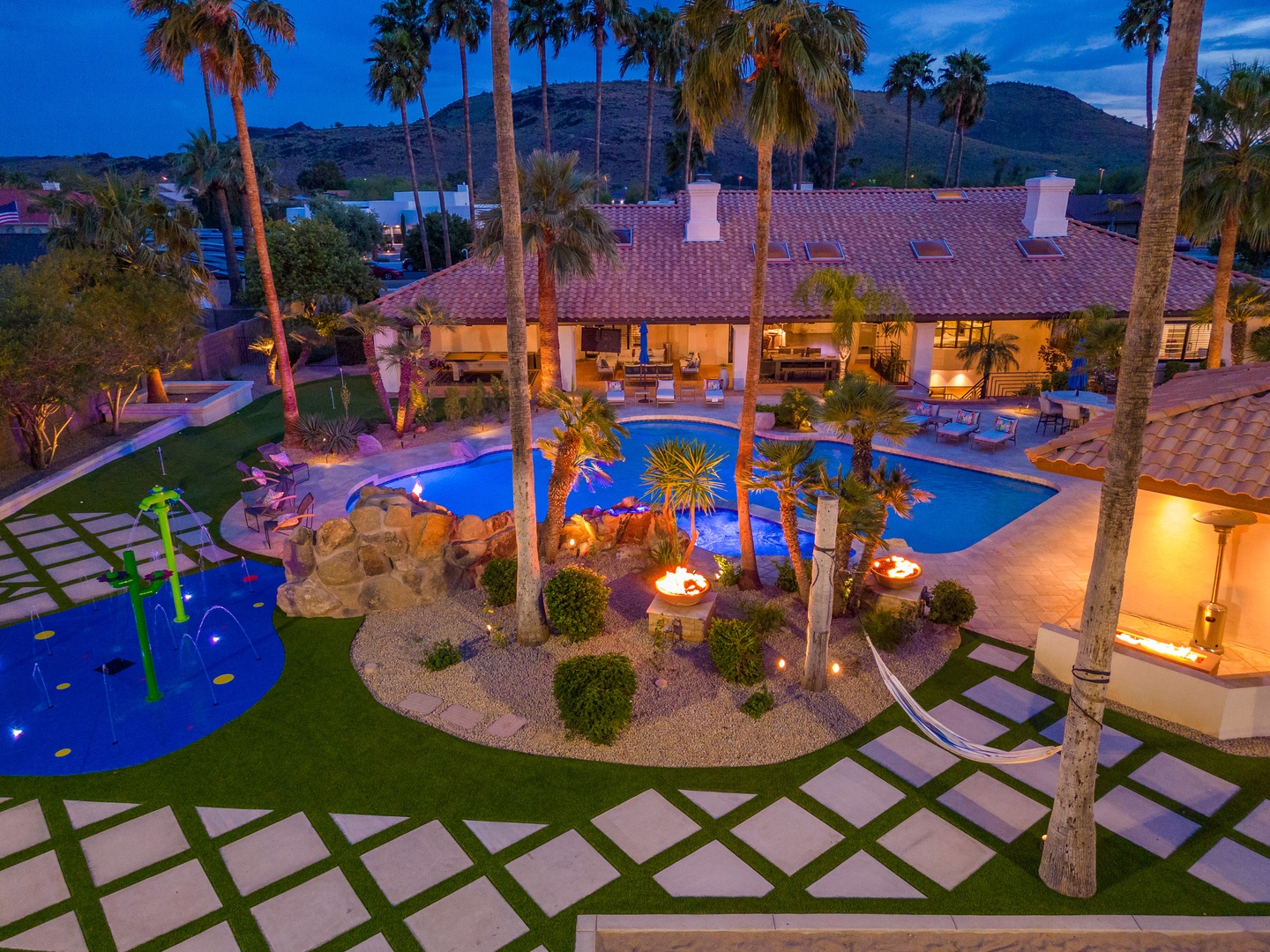 Aerial view of a luxurious vacation property featuring two pools, tropical palm landscaping, fire features, and mountain backdrop at dusk. The Spanish-style architecture and illuminated outdoor spaces create an inviting desert resort atmosphere.