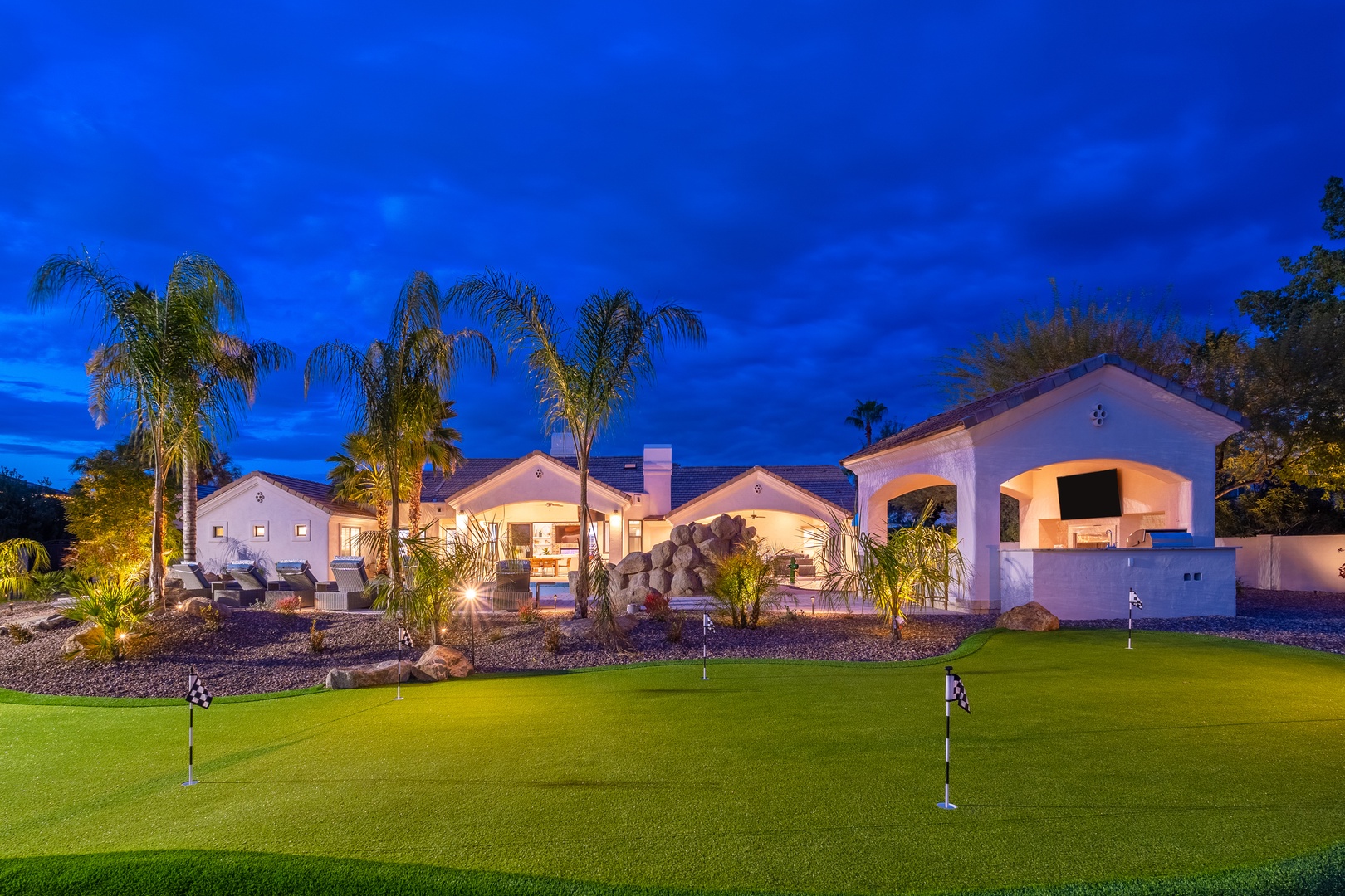 A stunning Spanish-style vacation home illuminated at twilight, featuring tropical palm landscaping and a private putting green in the front yard.