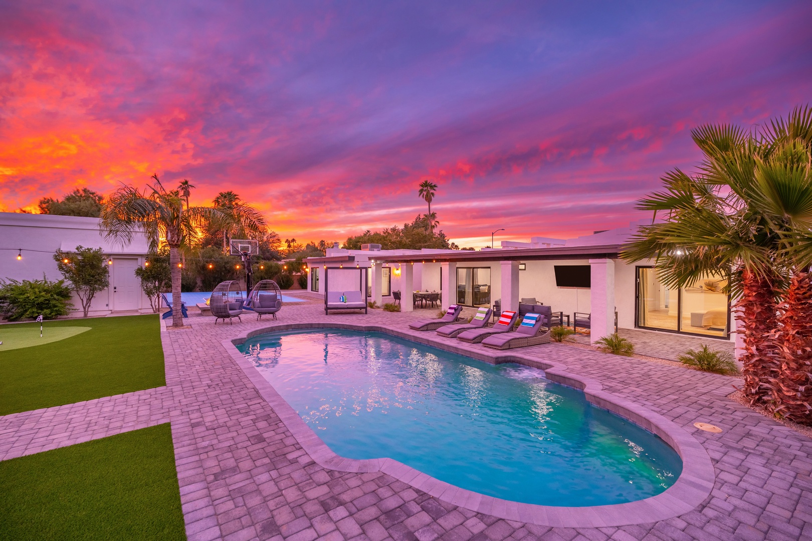 A stunning desert sunset paints the sky in brilliant pink and orange hues, creating a dramatic backdrop for this inviting property featuring a sparkling pool and palm-lined outdoor living spaces.