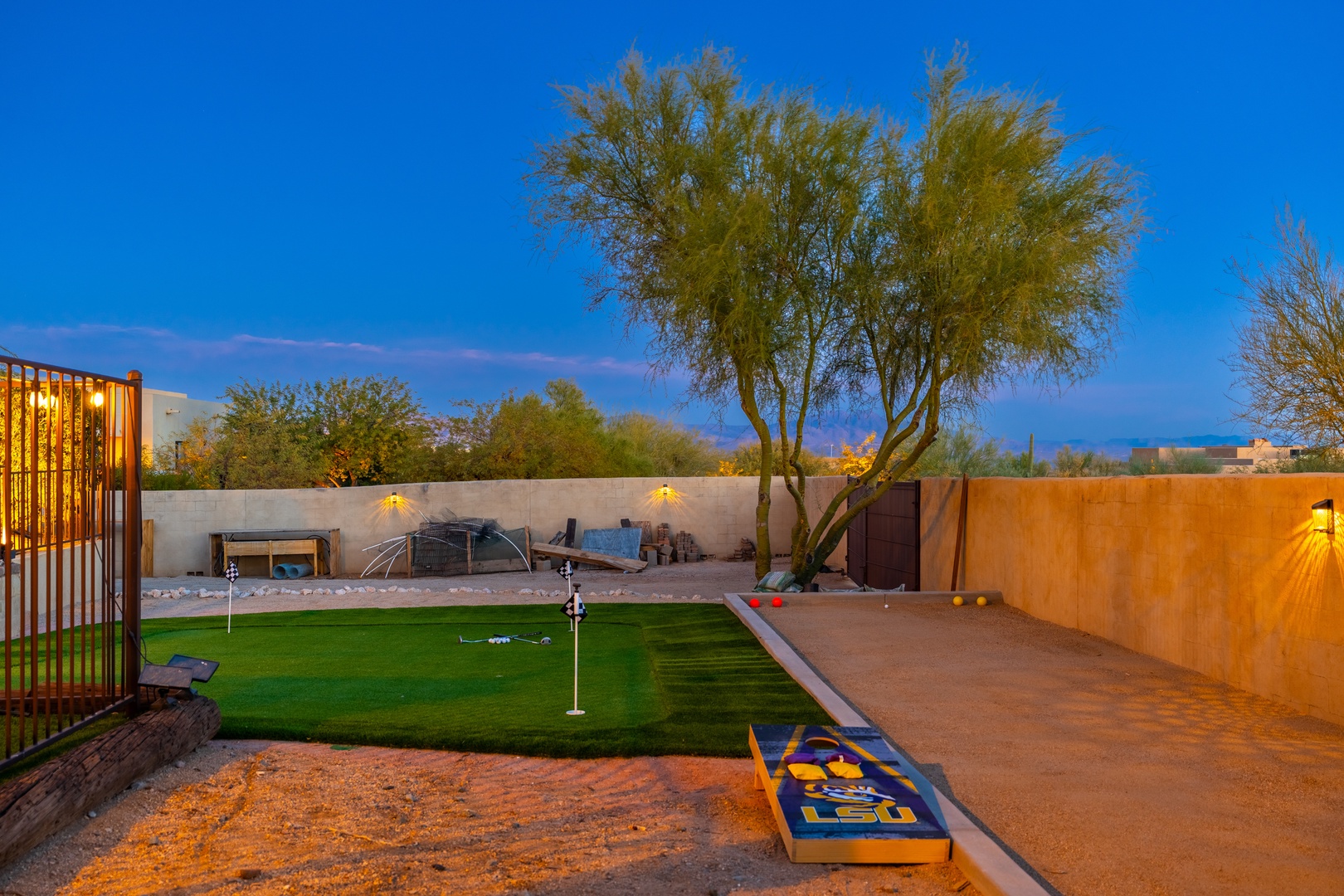 The backyard features a spacious lawn with putting green, cornhole, fire pit seating, and an outdoor lounge area—all framed by desert trees and mountain views under a clear blue evening sky.