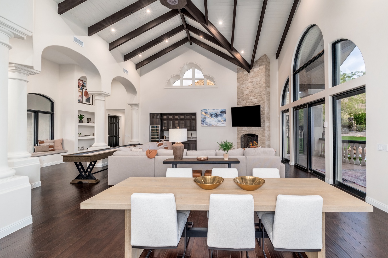 A stunning open-concept living space combines your dining, living, and kitchen areas under soaring vaulted ceilings with rustic exposed beams. Natural light pours through arched windows framing the cozy fireplace.