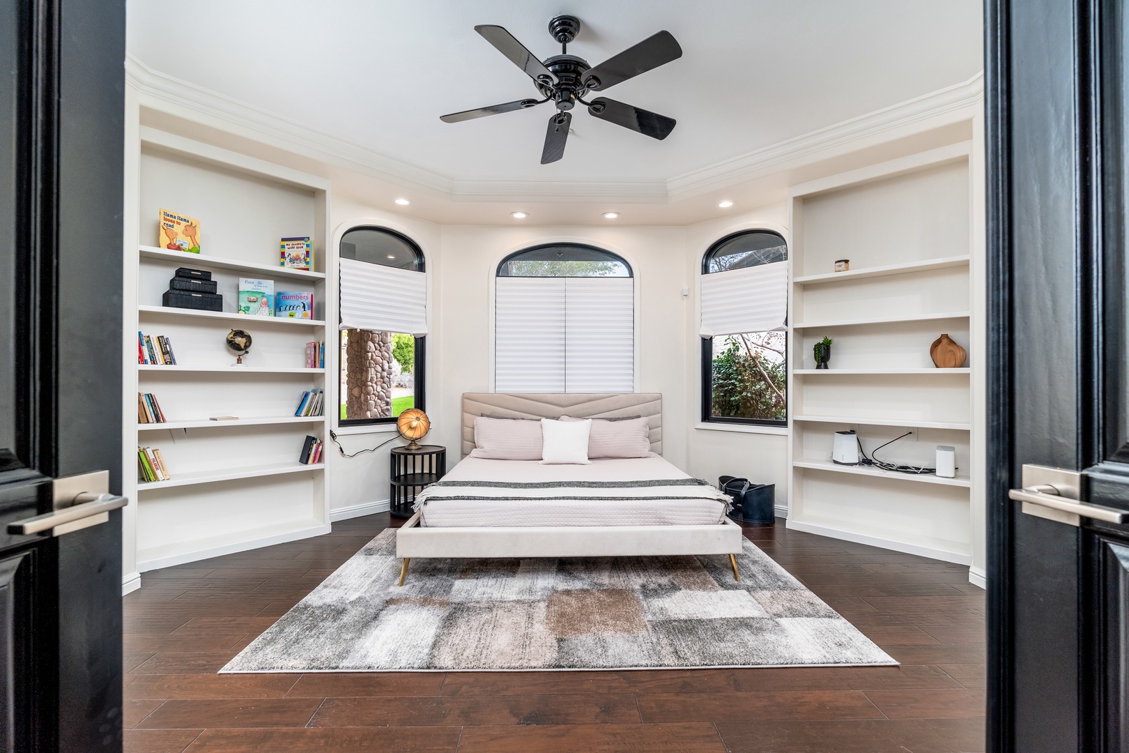 Settle into a bright bedroom framed by arched windows and built-in shelving. The ceiling fan keeps you cool while natural light filters through elegant window treatments.
