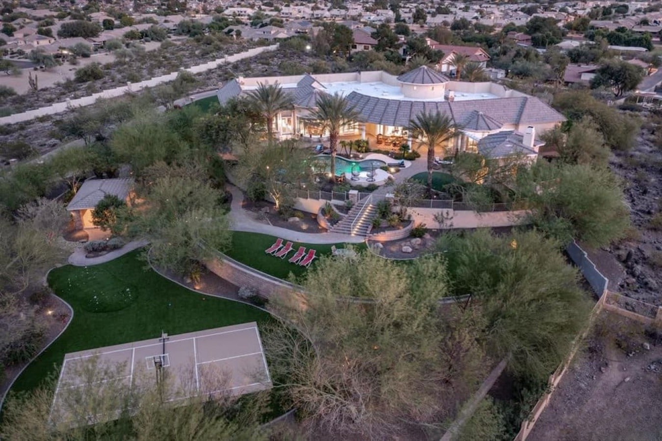 An expansive estate set within the desert landscape, featuring a main residence with palm trees, pool, tennis court, and manicured lawns surrounded by natural terrain.