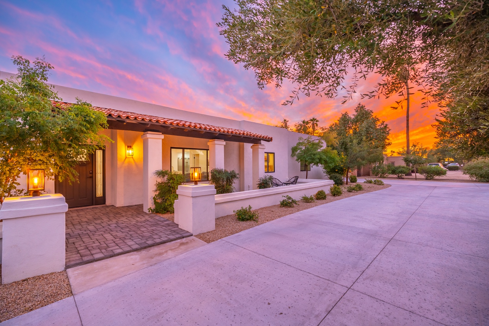 A stunning sunset paints the sky in vibrant pinks and oranges, framing the property's Spanish-style architecture. The inviting entrance features terracotta tile roofing, desert landscaping, and warm exterior lighting welcoming guests.