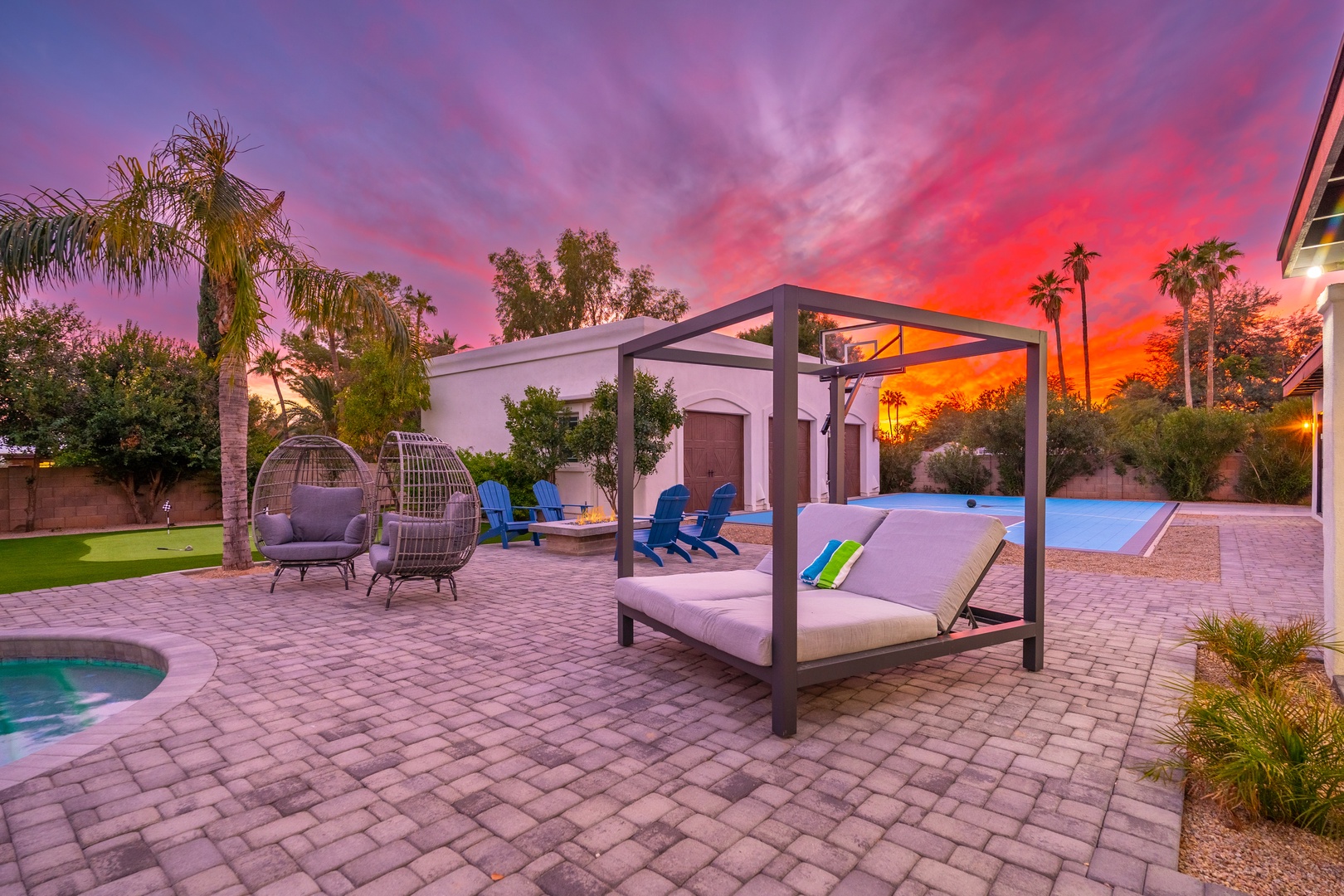 A stunning desert retreat features lush gardens, a sparkling pool, and an outdoor lounge beneath spectacular sunset skies painted in vibrant pinks and purples.