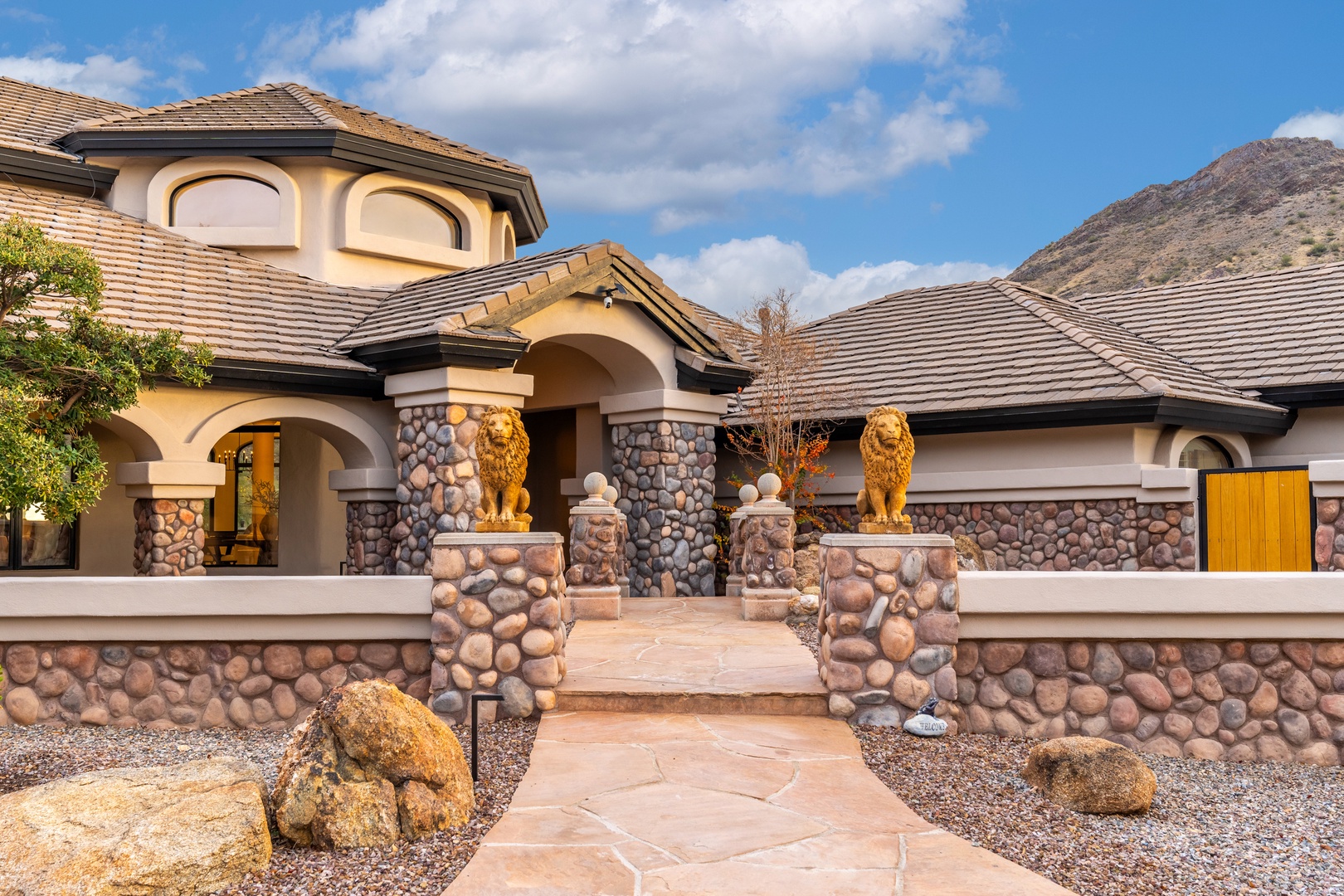 Elegant Mediterranean-style entrance showcases golden lion statues, stone archways, and mountain backdrop. A striking first impression for guests arriving at this desert retreat.