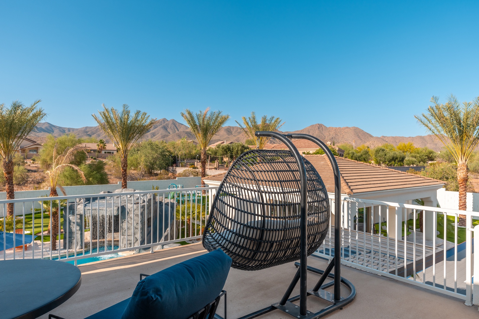 A desert oasis awaits beyond the balcony, where palm trees sway against mountain views and the pool glimmers below, inviting relaxation in your private hanging chair retreat.