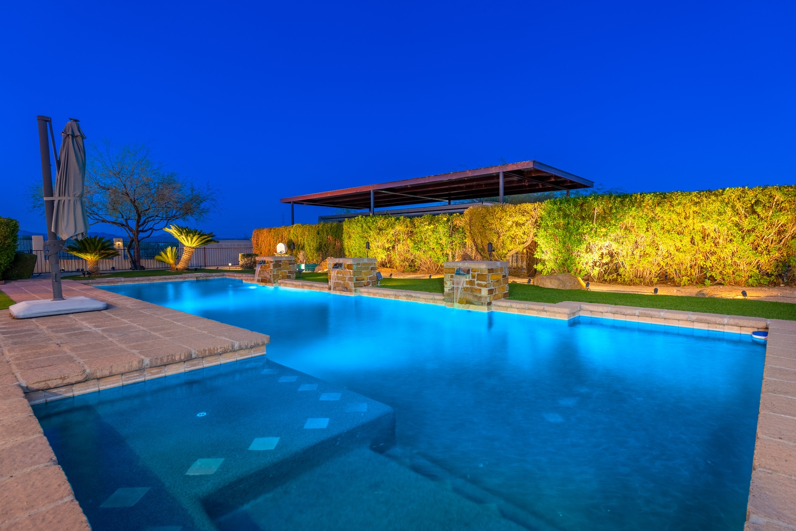 A stunning illuminated pool beckons at twilight, framed by lush greenery and a shaded lounge area perfect for evening relaxation.