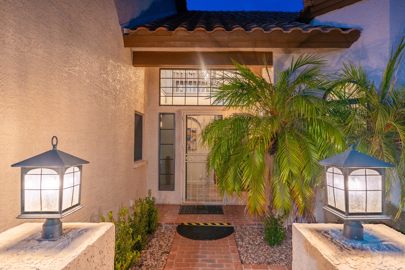 Charming property entrance showcases warm stucco walls, elegant lantern lighting, and swaying palm trees that frame the inviting front door at dusk.