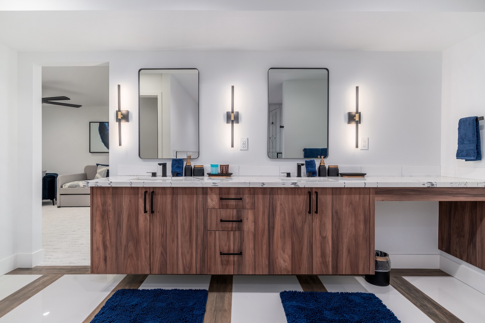 Your private retreat features a stunning double vanity with sleek marble countertops, warm walnut cabinetry, and modern fixtures. Elegant wall sconces illuminate your space, while plush blue accents add a touch of coastal charm.