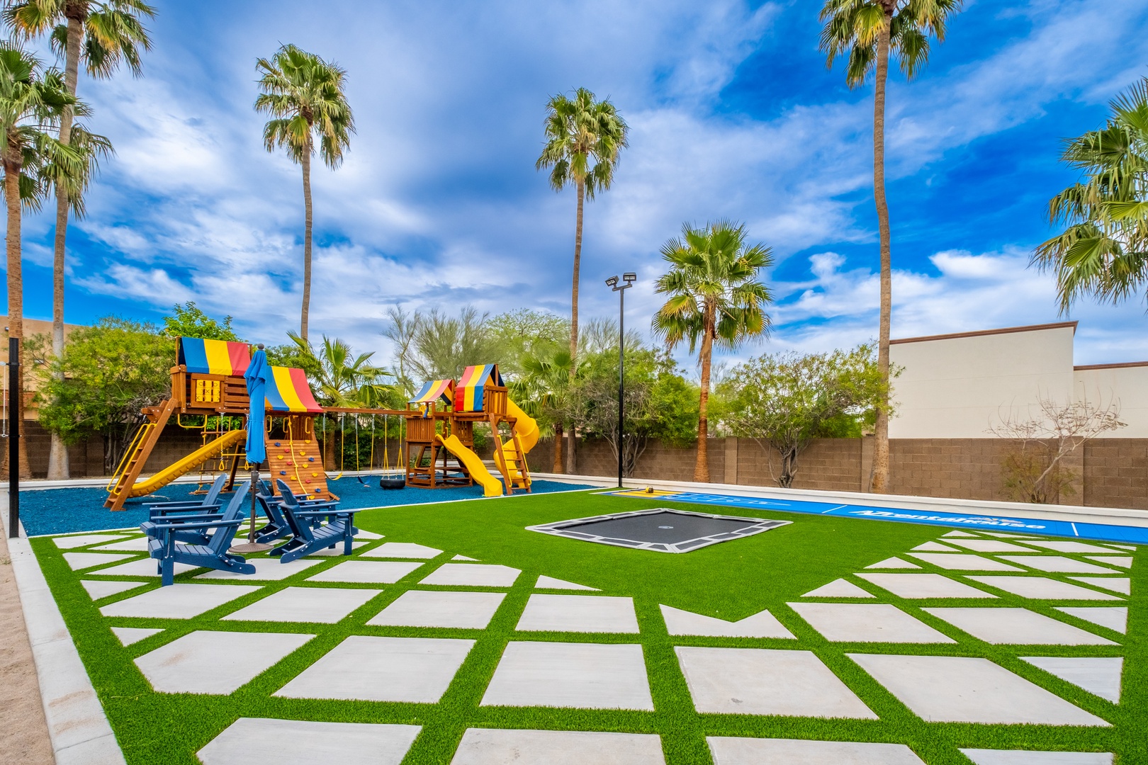Vibrant outdoor play area featuring a colorful playground, in-ground trampoline, and shaded palm trees—perfect for family fun under sunny skies.