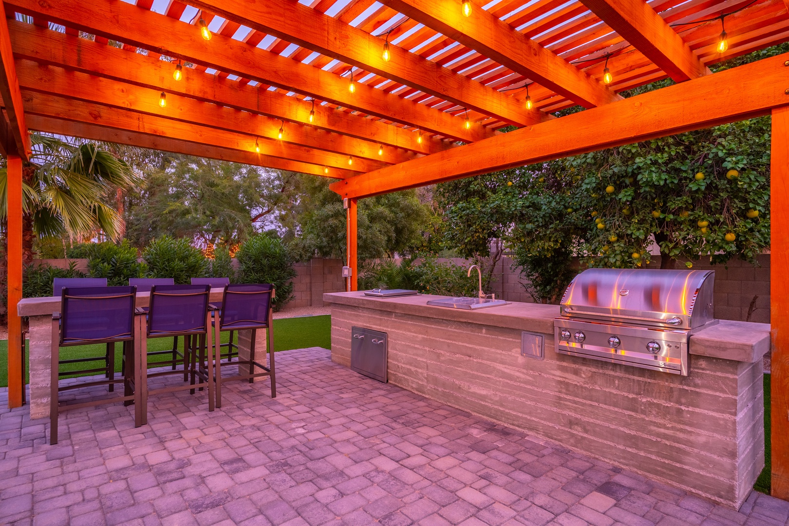A covered outdoor kitchen area with a built-in grill, sink, and bar seating, set against lush landscaping and string lighting overhead.