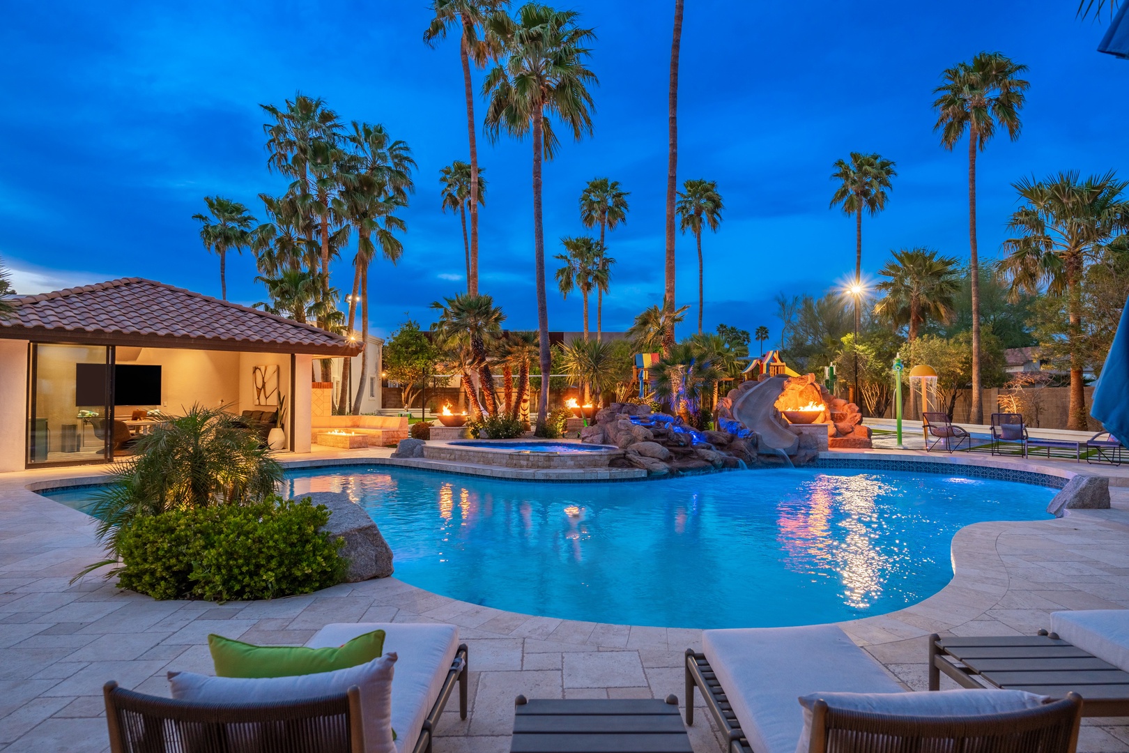A resort-style pool glows under dramatic twilight skies, framed by towering palms and illuminated rock features. The perfect spot for evening swims and relaxation.