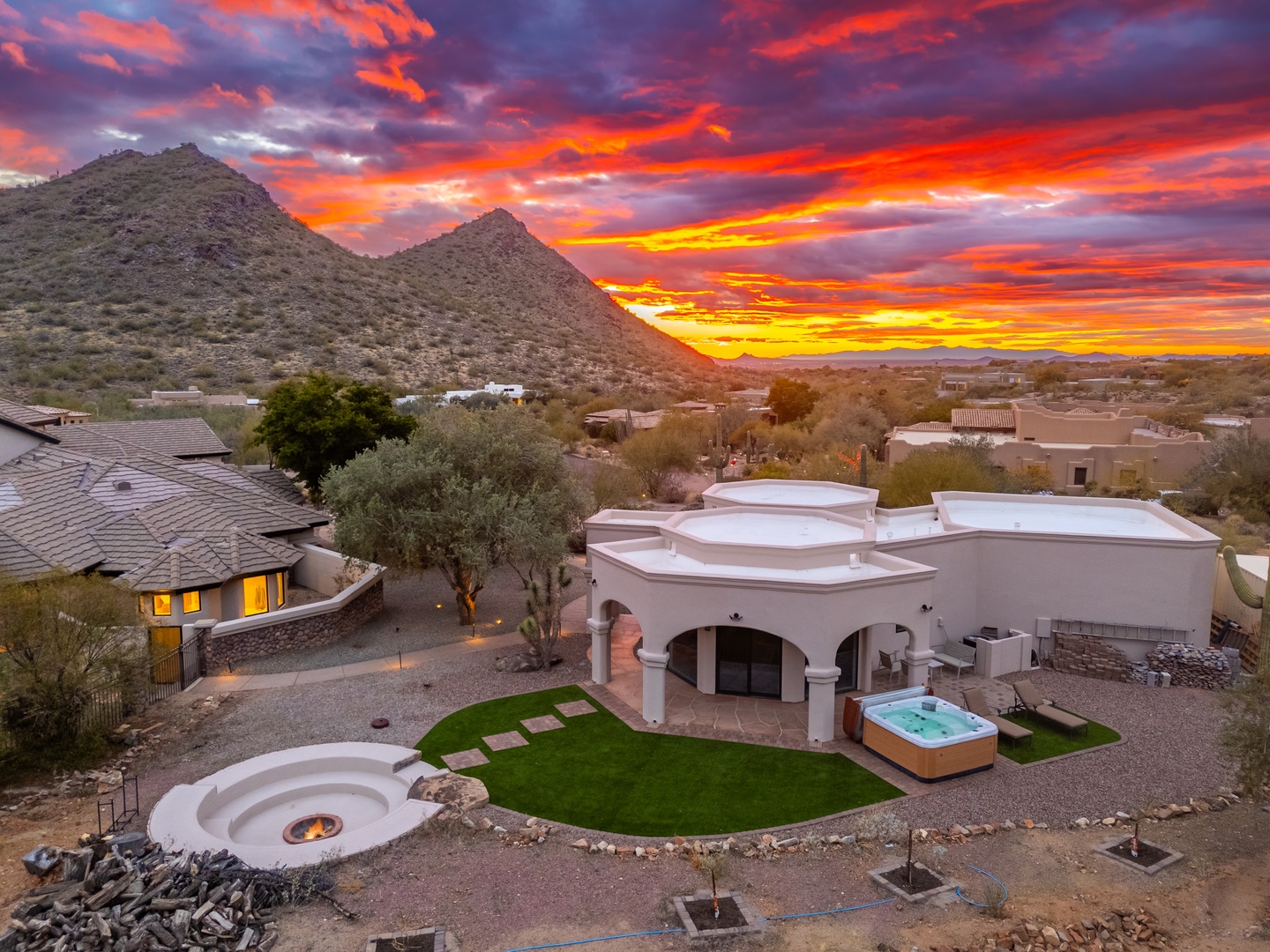 Aerial view of a Southwestern-style vacation home set against dramatic desert mountains, featuring a hot tub and landscaped yard, captured during a brilliant sunset painting the sky in vivid orange and purple hues.