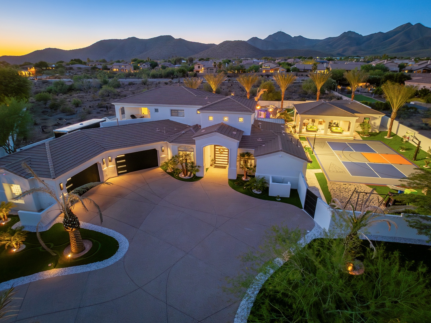 Expansive desert estate framed by dramatic mountain views and illuminated landscaping. Features private sports court, palm-lined grounds, and spacious outdoor living areas as evening falls.