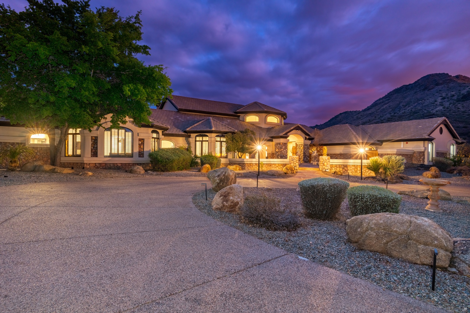 Expansive desert estate illuminated at twilight, framed by mountain backdrop and desert landscaping. The grand entrance and architectural details mark this property's distinctive presence in the natural terrain.