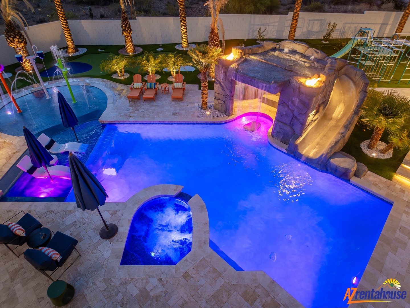 Stunning aerial view of this resort-style backyard oasis featuring a sparkling pool, relaxing spa, dramatic waterslide, and tropical palm trees illuminated beautifully at dusk.