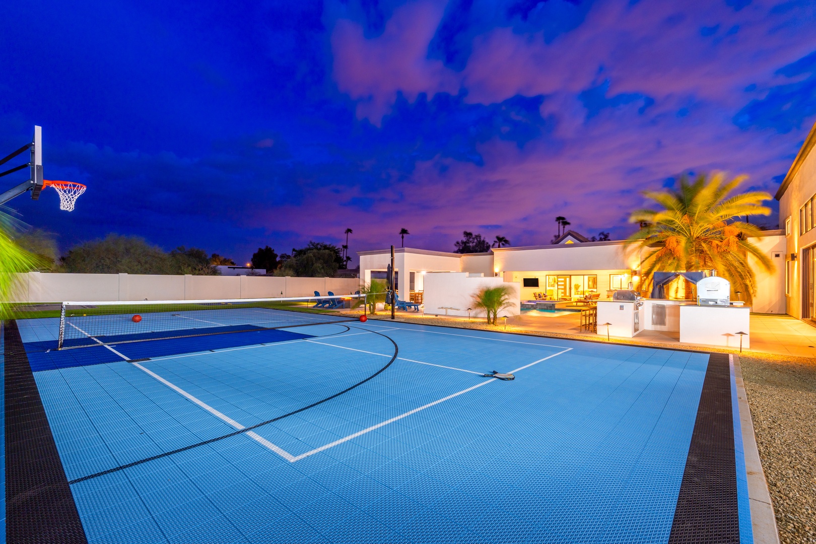 A private multi-sport court awaits under a stunning twilight sky, framed by palms and the inviting glow of the main house—perfect for evening play.