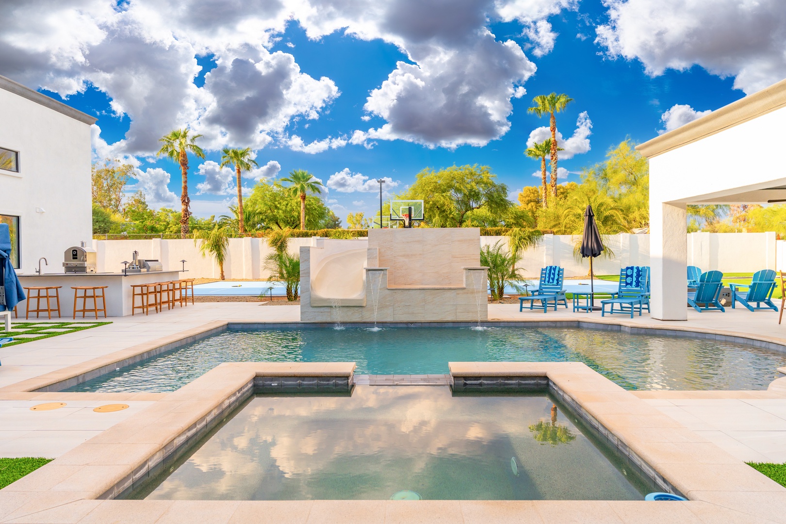 A resort-style backyard oasis awaits with pool, hot tub, basketball court, outdoor kitchen, and lush palm trees beneath stunning desert skies.