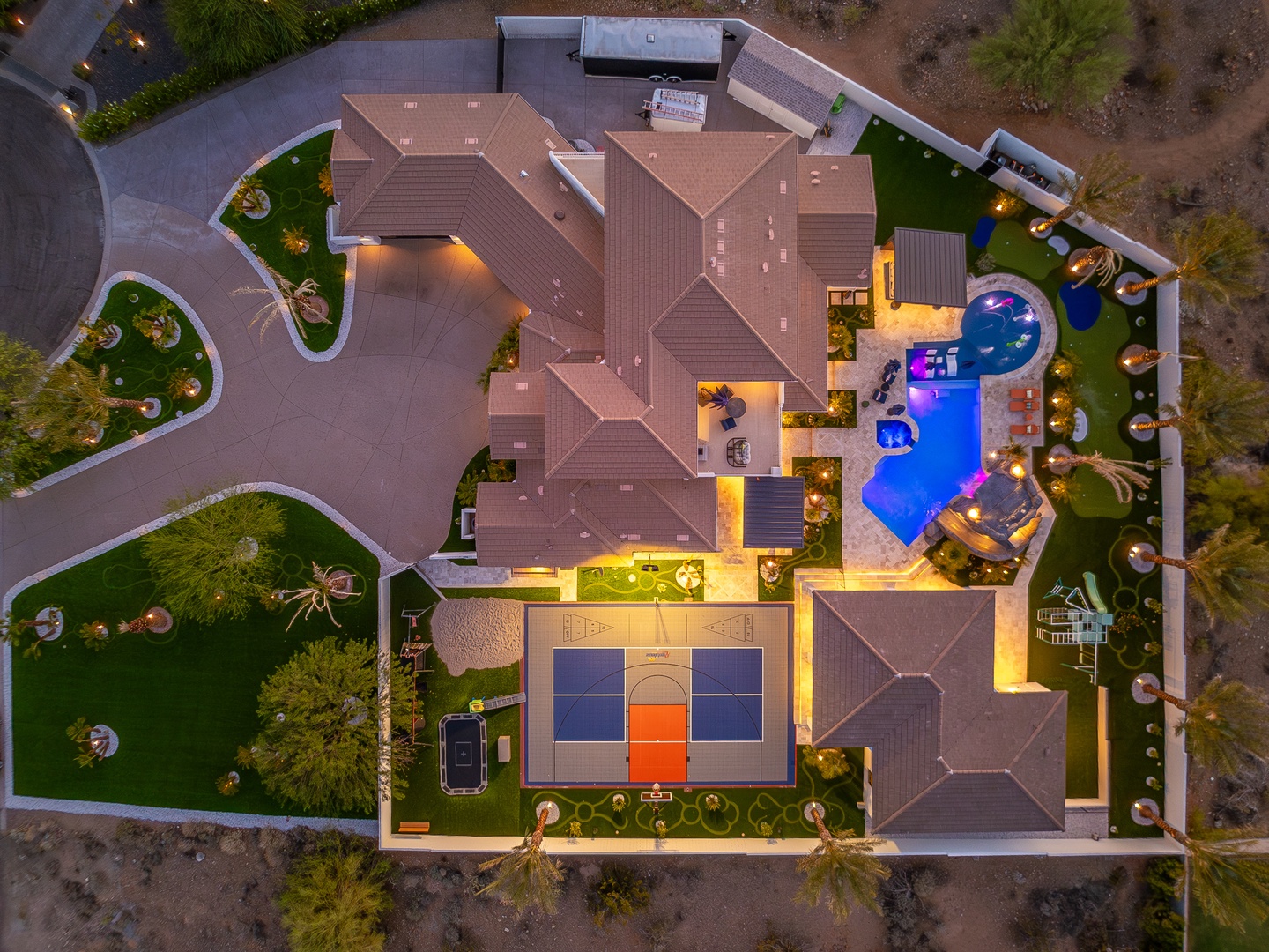 Stunning aerial view of the property showcasing a beautifully lit pool, sports court, lush gardens, and spacious outdoor entertaining areas—an impressive resort-style retreat.