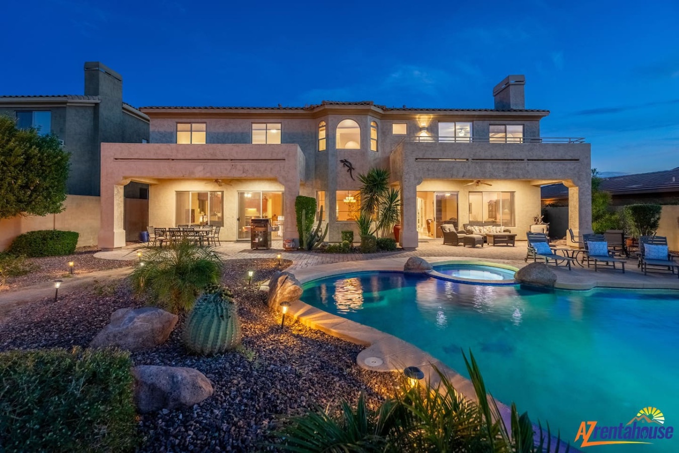 Luxury vacation home with sparkling pool, spa, and desert landscaping illuminated under evening skies.
