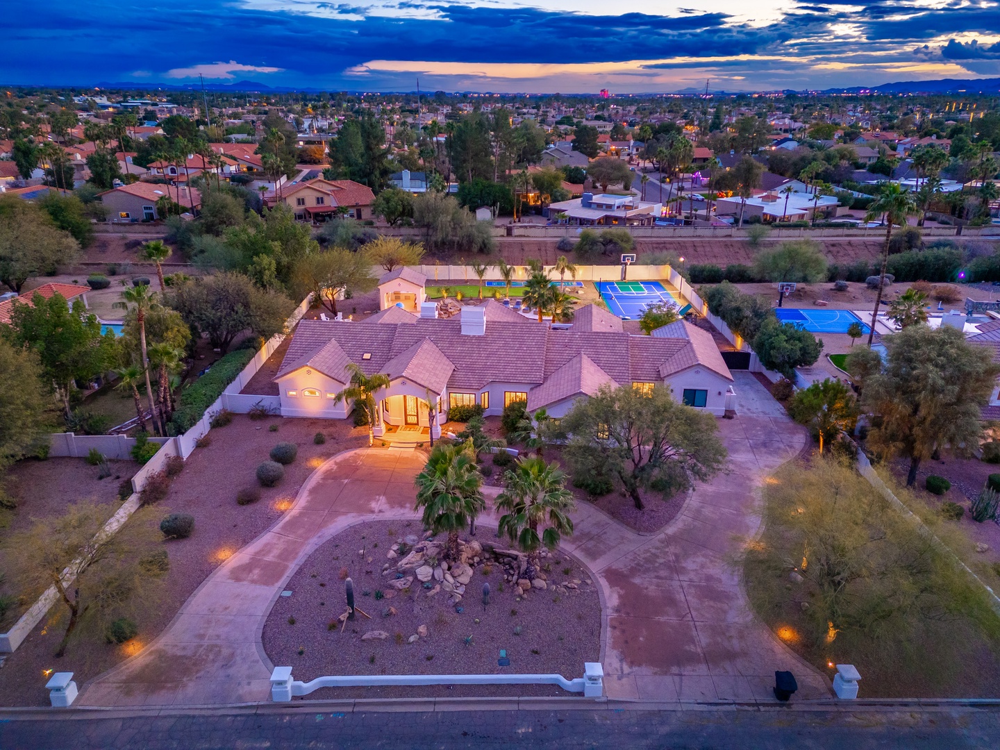 An aerial view captures this expansive vacation rental property at dusk, showcasing the main residence, pool area with sports courts, and surrounding neighborhood lighting up as evening settles in.