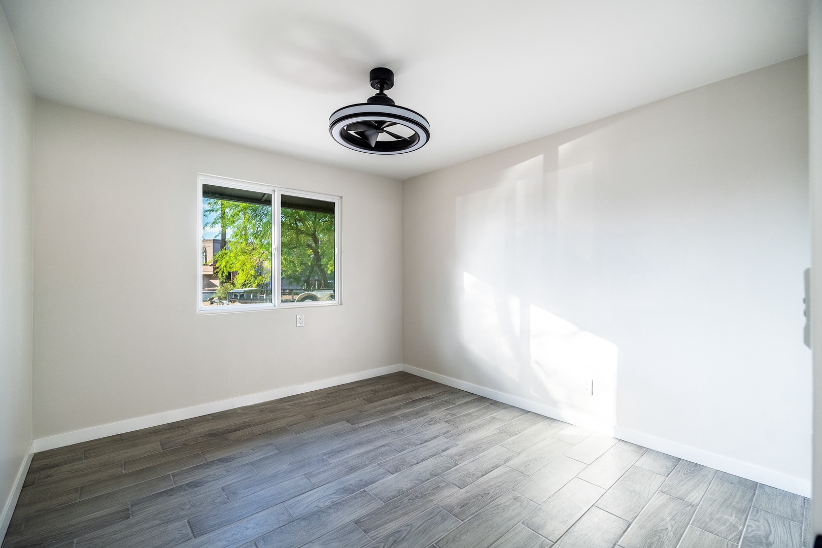 Step into your bright, empty-canvas bedroom with sleek wood-style floors, fresh white walls, and a modern ceiling fan ready to keep you cool. Sunlight streams through the window, inviting you to imagine your perfect retreat.
