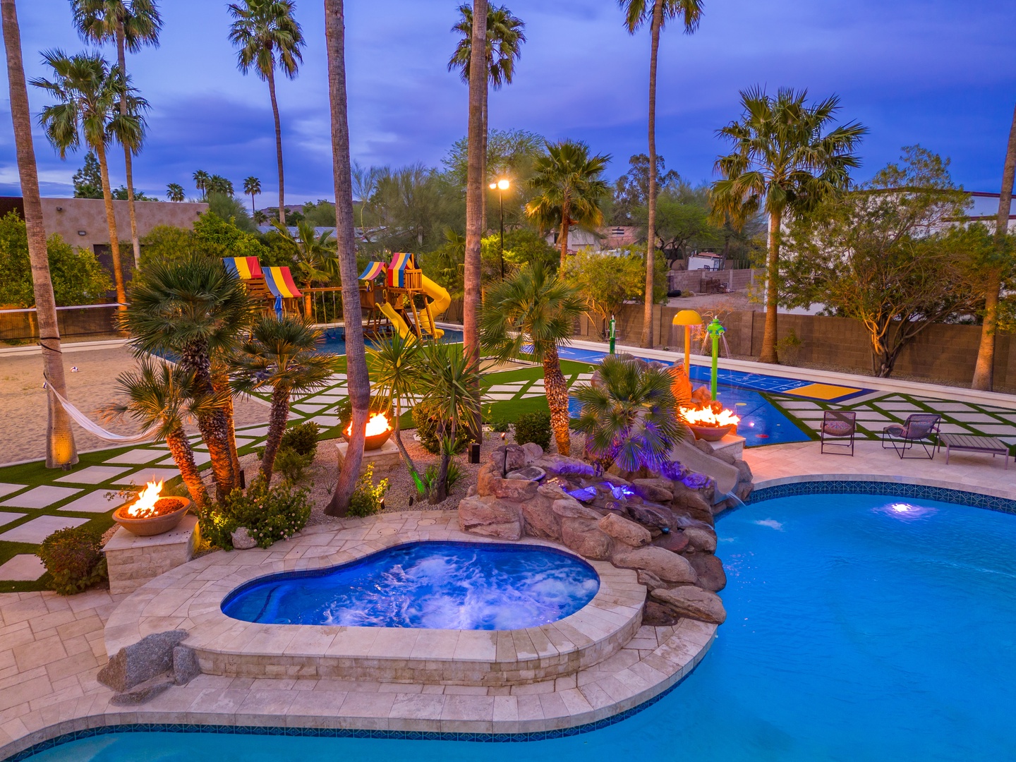 Resort-style backyard oasis captured at dusk, featuring a flowing pool with hot tub, surrounded by palm trees, ambient fire features, and a colorful children's play area beyond.