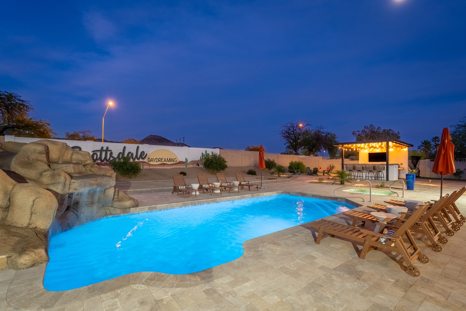 Evening pool area features a unique waterfall rock formation and glowing blue water, with lounge chairs and covered seating area nearby.