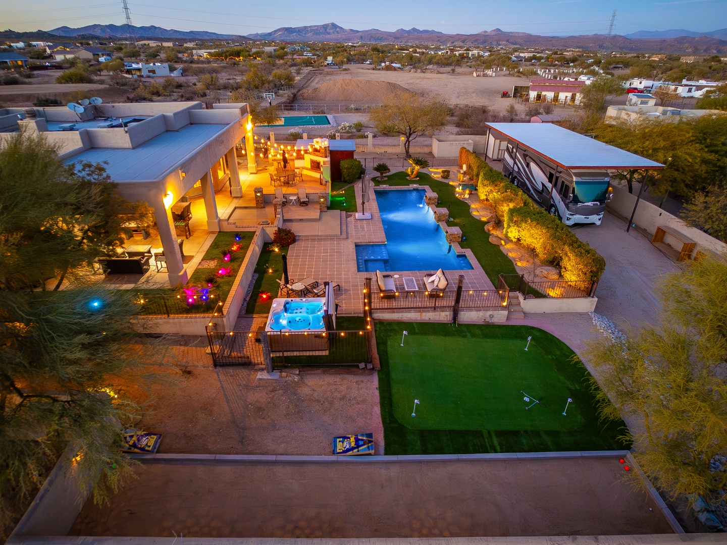 Aerial view showcasing the property's expansive outdoor amenities and desert mountain backdrop at twilight, featuring a large swimming pool, hot tub, putting green, and covered outdoor living areas.