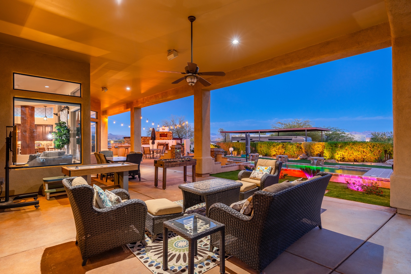 This covered patio offers stunning twilight views of the lush backyard, pool, and distant desert landscape. Relax in comfortable wicker seating, enjoy alfresco dining, or gather around the game table as day fades to night.