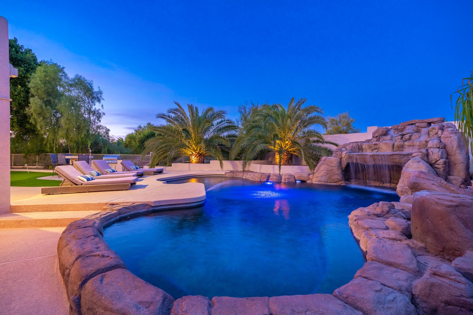 A stunning resort-style pool features dramatic rock formations, a cascading waterfall, and illuminated palms—perfect for evening relaxation under the open sky.