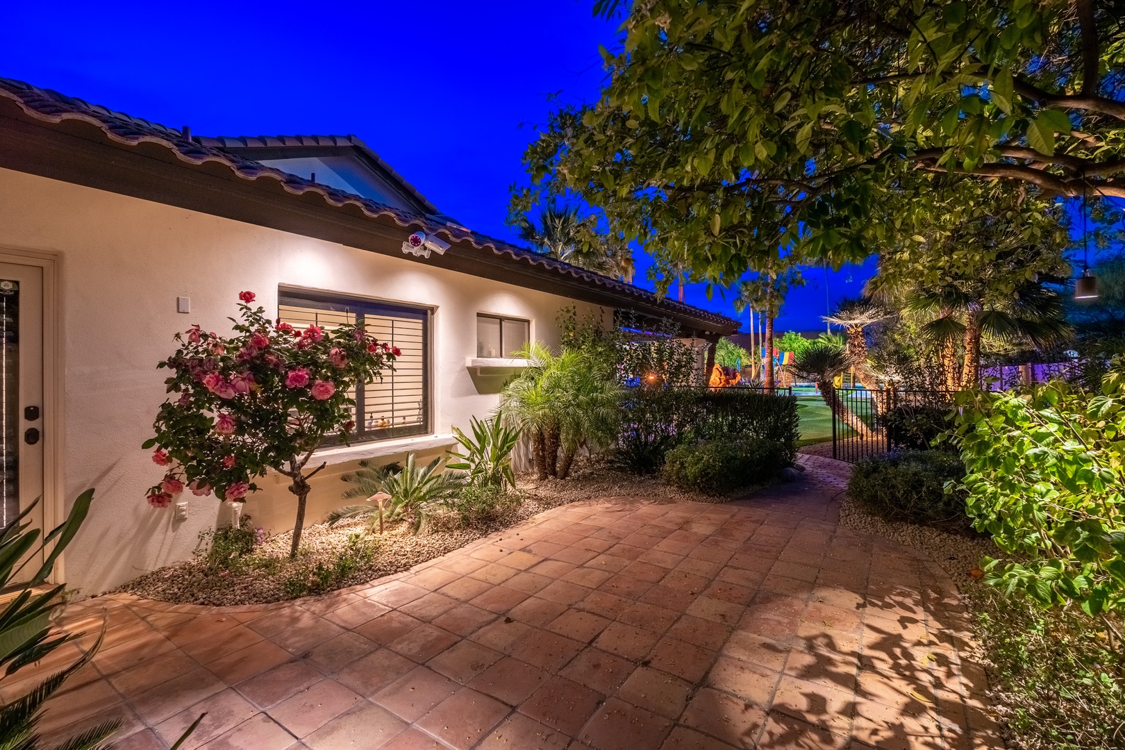 The property's exterior at twilight features stucco architecture with Spanish tile roofing, lush landscaping including flowering trees and palms, and paved walkways illuminated by ambient lighting.
