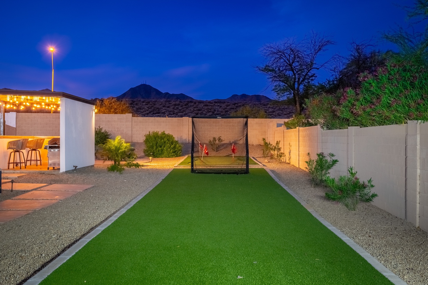 This property features a private outdoor recreation area with synthetic turf lawn and sports netting, surrounded by desert landscaping and mountain views, with an illuminated outdoor bar area.