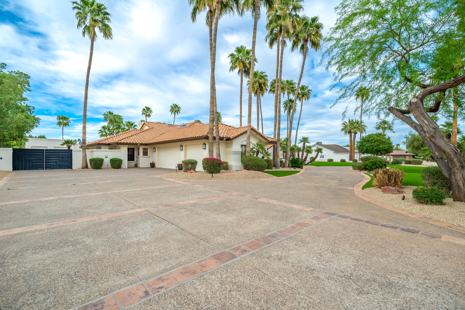 A single-story home with terracotta roof tiles sits beneath towering palms, surrounded by manicured desert landscaping and a spacious driveway with brick accent borders.