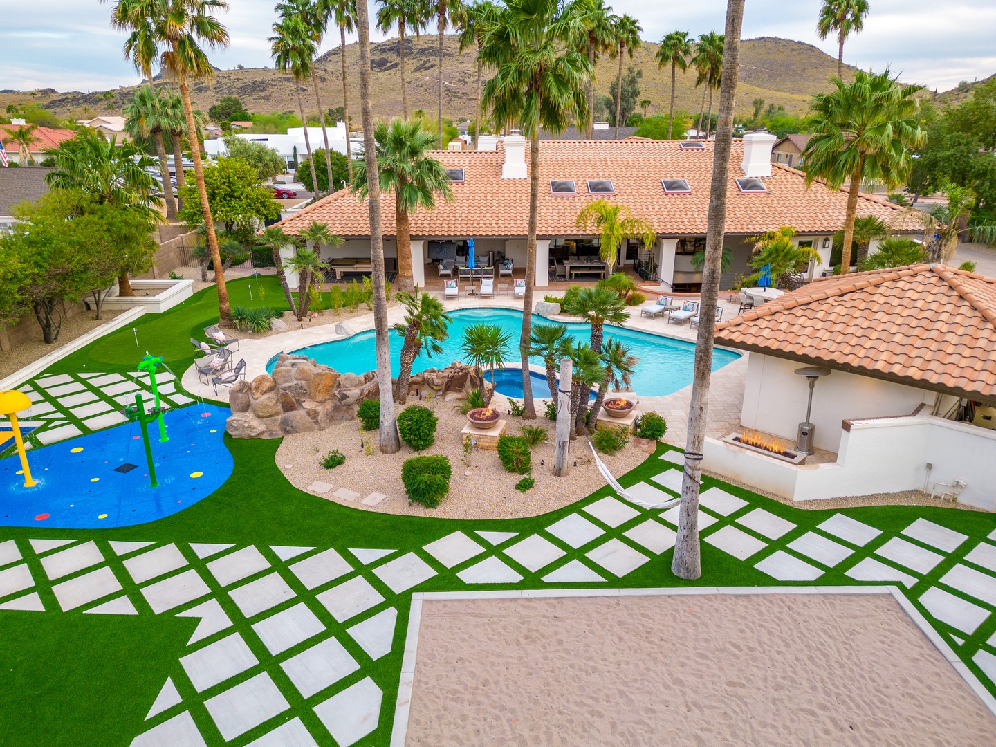 Aerial view of a desert oasis estate featuring a sparkling pool, palm-lined grounds, children's play area, and mountain backdrop—a resort-style property in a scenic hillside neighborhood.